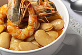 Schale mit typischem Bohneneintopf mit Garnelen, Krabben und Muscheln auf einer Spitzentischdecke neben einem Löffel und auf einem Steintisch