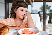 Wistful child with brown hair sitting at table with plates of delicious food in cafeteria and looking away