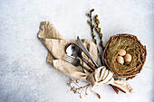 From above cutlery set for Easter dinner with dried flower twig and eggs on a bird nest on a concrete background