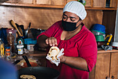 Mature ethnic female in cloth face mask cooking arepas with filling in house kitchen during COVID 19 pandemic