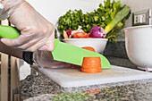 Crop anonymous female in gloves cutting slices of ripe orange carrot on chopping board with salad while preparing healthy vegetarian dish on counter with assorted vegetables in kitchen