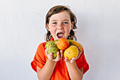 Adorable little boy in bright red shirt screaming happily and looking at camera while demonstrating assorted fresh exotic fruits against white background