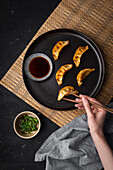 Top view of crop female with chopsticks taking gyoza from plate near bowls of soy sauce and greens during lunch on black table