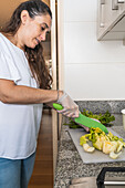 Side view of adult ethnic female cutting green fresh celery on cutting board using knife while cooking in kitchen at home