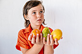 Adorable little girl in bright red shirt looking away while demonstrating assorted fresh exotic fruits against white background