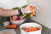Crop anonymous female in gloves throwing slices of ripe orange carrot in bowl with salad while preparing healthy vegetarian dish on counter with assorted vegetables in kitchen