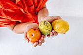 Cropped unrecognizable little girl in bright red shirt demonstrating assorted fresh exotic fruits against white background