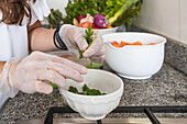 Crop anonymous female in gloves tearing fresh green parsley near bowls with ripe vegetables while cooking healthy vegetarian salad in kitchen at home