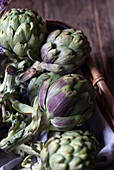 Closeup from above view of green artichokes laid in wicker basket with bunch of little purple flowers