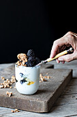 Glas voll mit Walnuss-Granola, gemischt mit frischen Blaubeeren und Joghurt, auf einem Holzbrett vor schwarzem Hintergrund