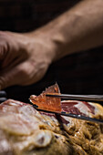 Crop unrecognizable cook cutting slice of tasty dry cured Spanish jamon on black background