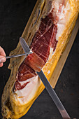 Draufsicht auf einen nicht erkennbaren Koch, der eine Scheibe leckeren, trocken gepökelten spanischen Jamon auf schwarzem Hintergrund schneidet