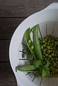 Top view of white plastic drain bowl full of peas and pods and fresh mint leaves on wooden table