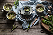 Flache Schalen mit grüner Erbsen- und Kokosnusscremesuppe auf Holztisch mit Erbsenschoten und Knoblauch in Komposition