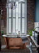 Freestanding copper bathtub in front of window with shutters and brick wall