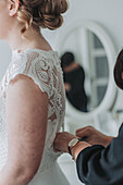 Braut in Spitzenhochzeitskleid vor Spiegel erhält Hilfe beim Ankleiden