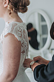 Bride in lace wedding dress in front of mirror receiving help with dressing