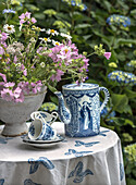 Blumentopf mit Sommerblumen und blau-weißes Teeservice auf Tischdecke mit Schmetterlingen