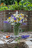 Blumenstrauß aus Rittersporn (Delphinium), Färberkamille (Anthemis tinctoria), Etagenzwiebel, Große Knorpelmöhre, auf Gartenmauer im Bierkrug