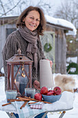 Frau mit Laterne und Keramik Krug, Tasse, Äpfel, Zimtstangen in winterlichem Garten und Gartenhütte