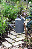 Garden path made of light-coloured paving stones with grey watering can and flower beds