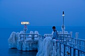 Schweiz, Kanton Waadt, Versoix, die Ufer des Genfer Sees sind bei sehr kaltem Wetter mit Eis bedeckt, eine Schaufensterpuppe auf dem mit Eis bedeckten Steg in der Abenddämmerung