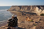 Marokko, Westsahara, Dakhla, alter Mann, der auf einer Klippe sitzt und den Strand von Araiche und seine Fischerboote überblickt