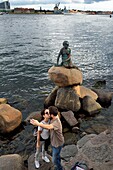 Denmark, Zealand, Copenhagen, couple of tourists in love in front of the Little Mermaid on its rock, bronze statue of Edvard Eriksen