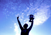 Silhouette of man holding up trophy against sky