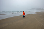 Woman in orange jacket walks along foggy beach