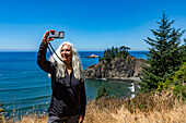 USA, Oregon, Brookings, Ältere Frau macht Selfie mit Küstenlandschaft im Hintergrund