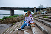 Nachdenkliche Frau sitzt auf den Stufen und trinkt Kaffee, während sie auf den Fluss schaut