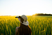 Rückansicht einer Frau mit Strohhut, die bei Sonnenuntergang in einem Feld steht