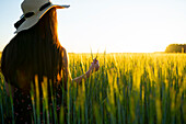Rückansicht einer Frau mit Strohhut, die bei Sonnenuntergang in einem Feld steht