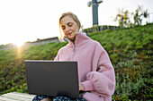 Frau arbeitet an einem Laptop, während sie bei Sonnenaufgang auf einer Bank sitzt