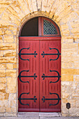 Saintes-Maries-de-la-Mer, Bouches-du-Rhone, Provence-Alpes-Cote d'Azur, Frankreich. Rote Tür mit Metallscharnieren in einem Steingebäude.