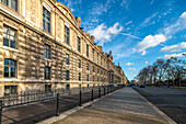 Die Fassade des Louvre erstrahlt im Sonnenlicht bei blauem Himmel und einem von Bäumen gesäumten Weg.