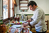 Side view of focused male chef in uniform cutting onion on board while standing at table with assorted ingredients in kitchen