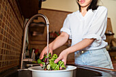 Anonymous female washing fresh lettuce leaves in colander under running water while cooking healthy food in kitchen at home