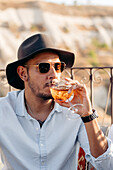 Young bearded male in sunglasses and stylish hat sipping cold refreshing cocktail while relaxing on terrace in Cappadocia, Turkey