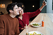 Fröhliches, junges, gemischtrassiges Paar sitzt am Tresen mit gesundem vegetarischem Salat und Getränken und macht ein Selfie mit dem Smartphone beim gemeinsamen Frühstück in einem modernen Restaurant