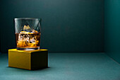 Glass cup with cold whiskey and cubes of ice placed on a gold-colored rugged base and a turquoise blue corner background
