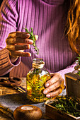 Crop faceless female in purple sweater putting herbs twigs with green leaves in essential oil glass bottle near scissors and rope with small chest on cloth at wooden table
