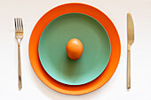 From above of raw brown chicken egg placed on colored ceramic plates served on white table with silver fork and knife