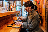 Seitenansicht einer zufriedenen asiatischen Frau im Pullover, die lächelnd einen Löffel von einem Mitarbeiter nimmt, der an einem Holztresen in einer Ramen-Bar sitzt