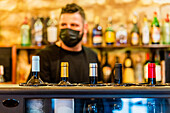 Unscharfer Barmann mit steriler Maske steht am Bartresen, während er in einem Restaurant während des Coronavirus arbeitet