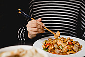 Unrecognizable female using chopsticks to eat portion of delicious Gong Bao chicken against black background in restaurant