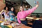 Happy young female friends in casual clothes taking selfie on mobile phone while having lunch together in modern restaurant