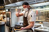 Male cook in uniform and mask adding pinch of seasoning on pan with meal on wooden board in modern restaurant during coronavirus pandemic