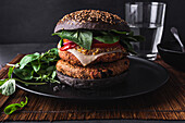 Appetitlicher Burger mit Sesam auf schwarzem Teller neben Salatblättern vor einem Glas Wasser auf dunklem Hintergrund